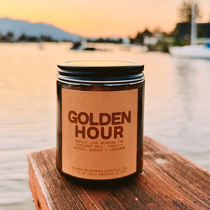 Candle jar labeled 'Golden Hour' on a wooden dock with a sunset over water background