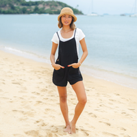Woman in black overalls and straw hat standing on a beach with ocean in the background