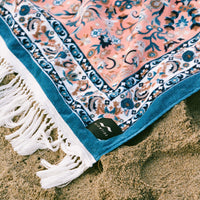 Decorative towel with floral pattern and tassels on sand, featuring a visible brand label.
