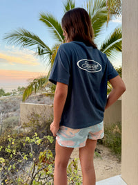 Person wearing a navy blue shirt with a logo, standing in a tropical setting with palm trees and sunset.