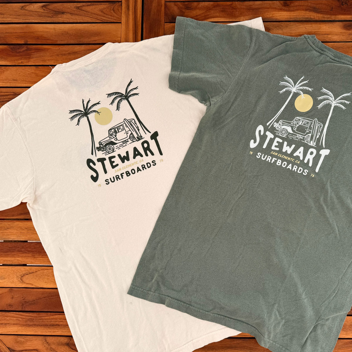 Two t-shirts with 'Stewart Surfboards' logos on a wooden surface