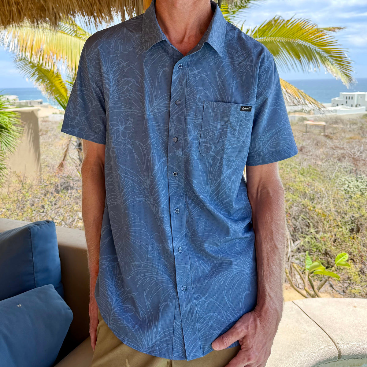man wearing a blue button-up shirt with palm trees and ocean in the background