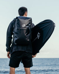 A man seen from behind standing at the beach, wearing a black waterproof backpack and holding a large black surfboard case.