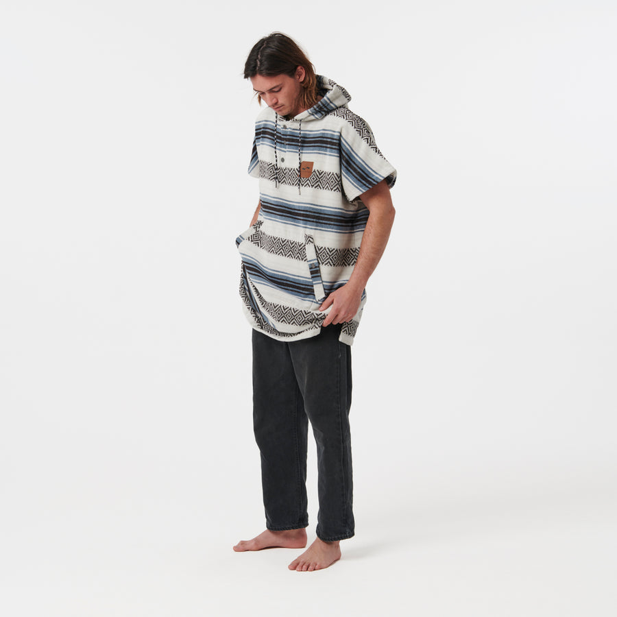 Person wearing a patterned poncho and dark pants on a white background