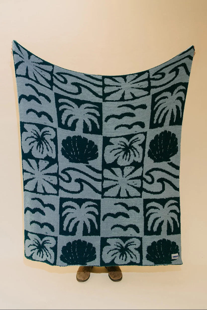 A rectangular woven blanket with a tropical pattern of palm trees, hibiscus flowers, and waves in dark teal and light blue, held up against a plain beige wall.