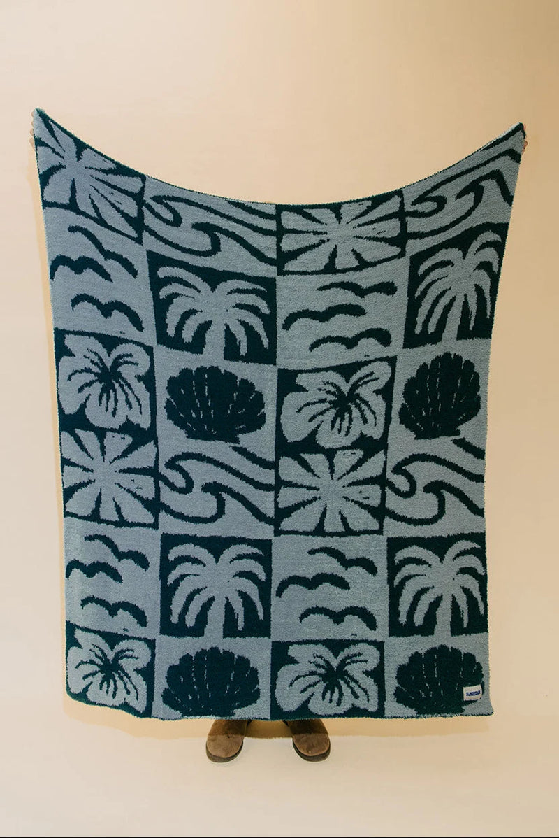A rectangular woven blanket with a tropical pattern of palm trees, hibiscus flowers, and waves in dark teal and light blue, held up against a plain beige wall.