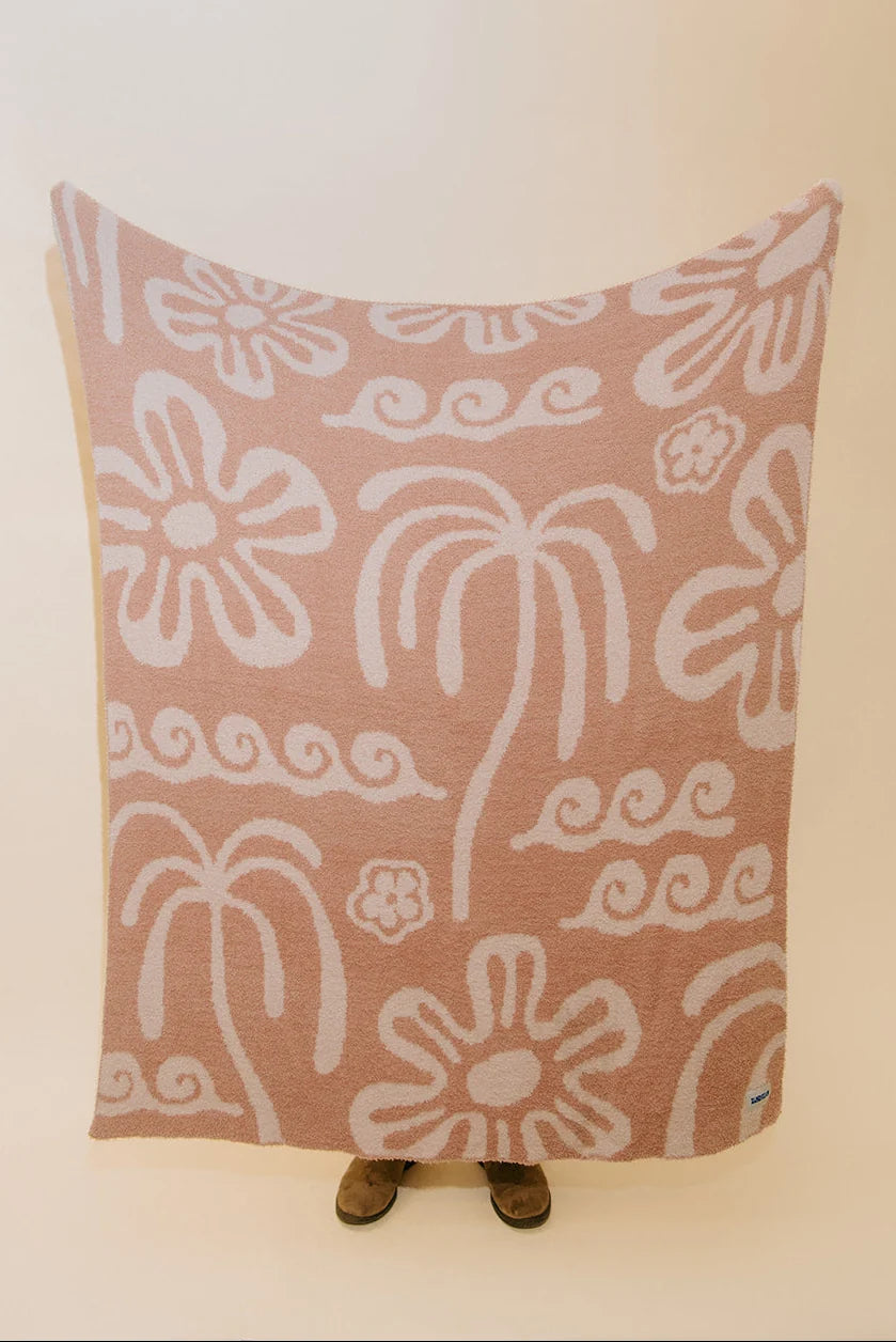 A large rectangular textile blanket or throw with a dusty pink background and white hand-drawn style floral and scroll motifs, held up against a plain light wall. Small feet are visible underneath the blanket, and a tiny blue tag appears at the lower right corner.