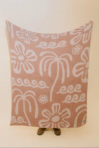 A large rectangular textile blanket or throw with a dusty pink background and white hand-drawn style floral and scroll motifs, held up against a plain light wall. Small feet are visible underneath the blanket, and a tiny blue tag appears at the lower right corner.