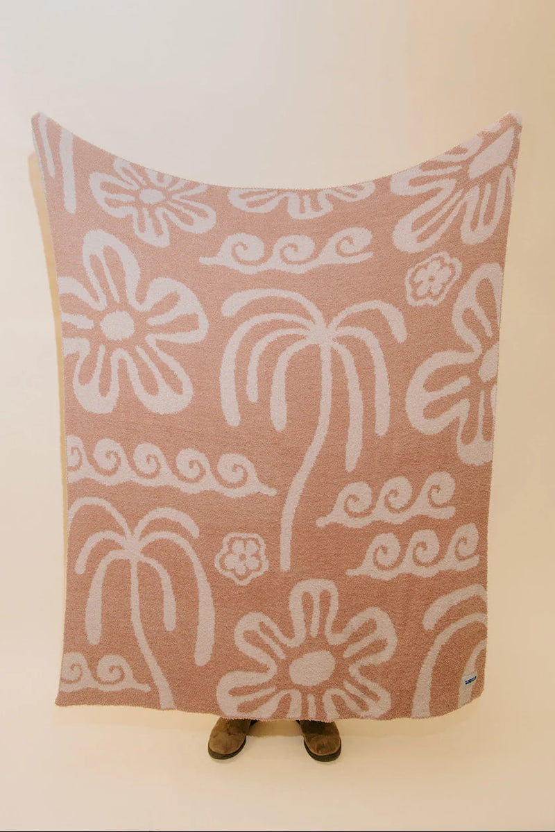 A large rectangular textile blanket or throw with a dusty pink background and white hand-drawn style floral and scroll motifs, held up against a plain light wall. Small feet are visible underneath the blanket, and a tiny blue tag appears at the lower right corner.