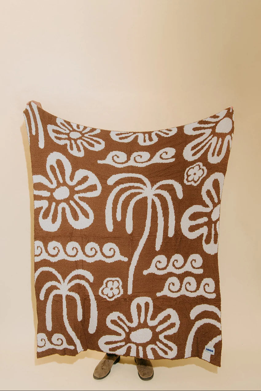 A brown textile blanket or throw with a white hand-drawn style floral and palm tree pattern, draped against a plain beige wall with a person's feet visible at the bottom.