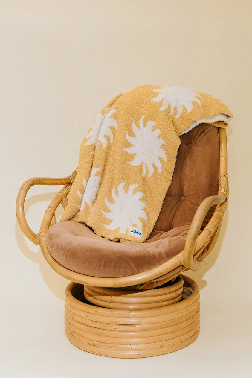 A mustard yellow plush throw blanket with a white sunburst pattern draped over a rattan swivel chair with brown velvet cushions, set against a plain beige background.