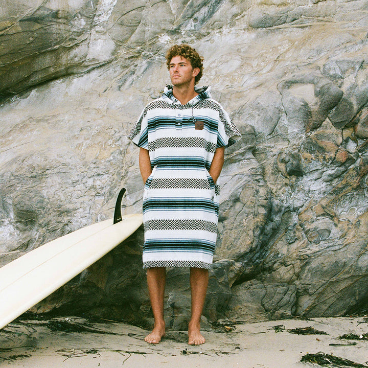 Man standing on a rocky beach wearing a striped towel, with a surfboard beside him.