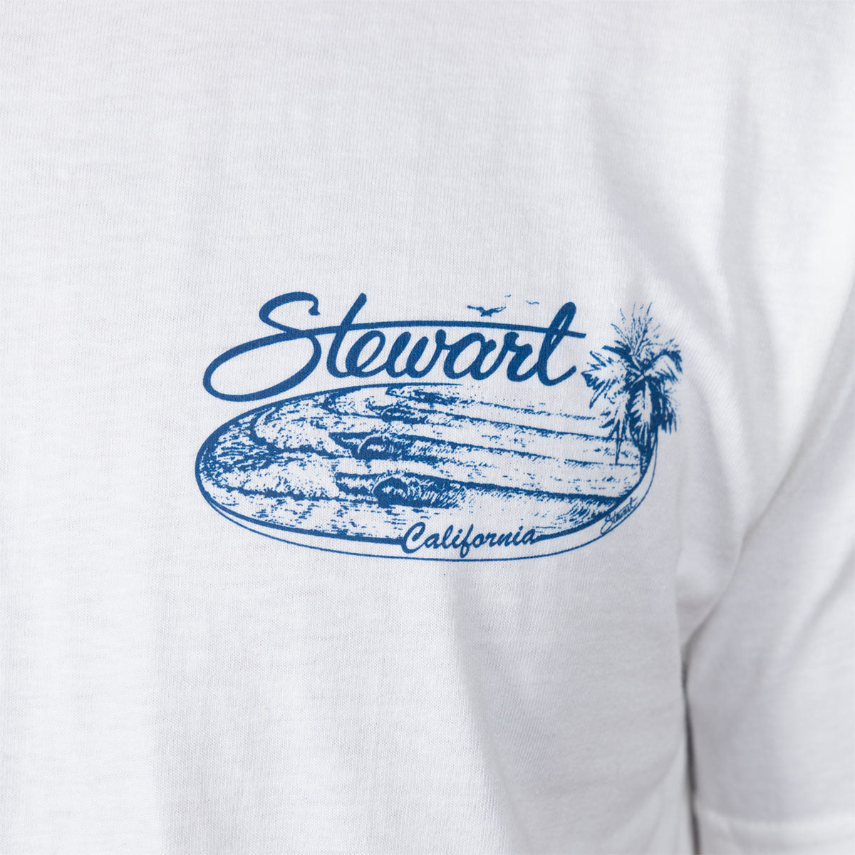 Close-up of a white T-shirt with a blue graphic print reading 'Stewart California' over a surfboard and palm tree design.