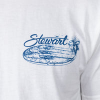 Close-up of a white T-shirt with a blue graphic print reading 'Stewart California' over a surfboard and palm tree design.
