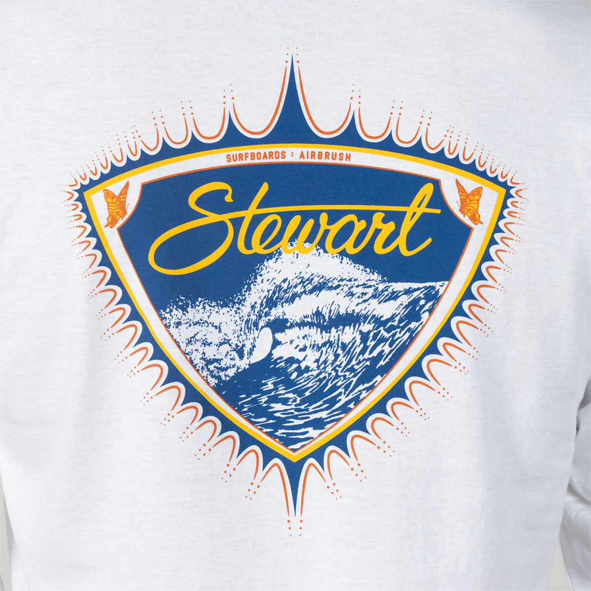 A close-up of a white long-sleeve shirt featuring a large vintage-style graphic on the chest. The graphic is a shield shape with a blue background, yellow 'Stewart' script, white surfboard and wave illustration, and orange and blue decorative border details.