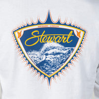 A close-up of a white long-sleeve shirt featuring a large vintage-style graphic on the chest. The graphic is a shield shape with a blue background, yellow 'Stewart' script, white surfboard and wave illustration, and orange and blue decorative border details.