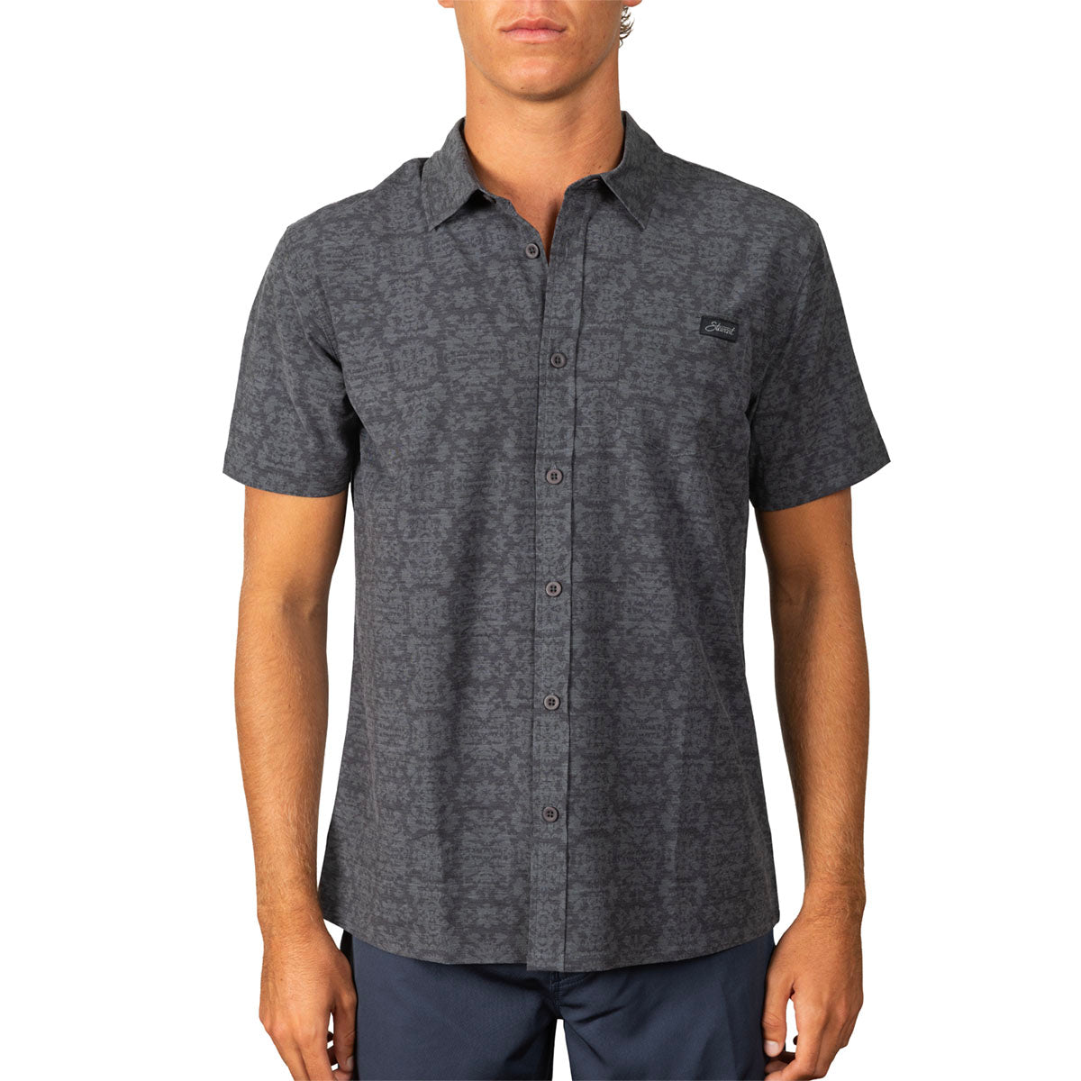 A man wearing a dark gray short-sleeve button-up shirt with a subtle tonal pattern and a small black logo patch on the left chest, paired with navy pants, against a plain white background.