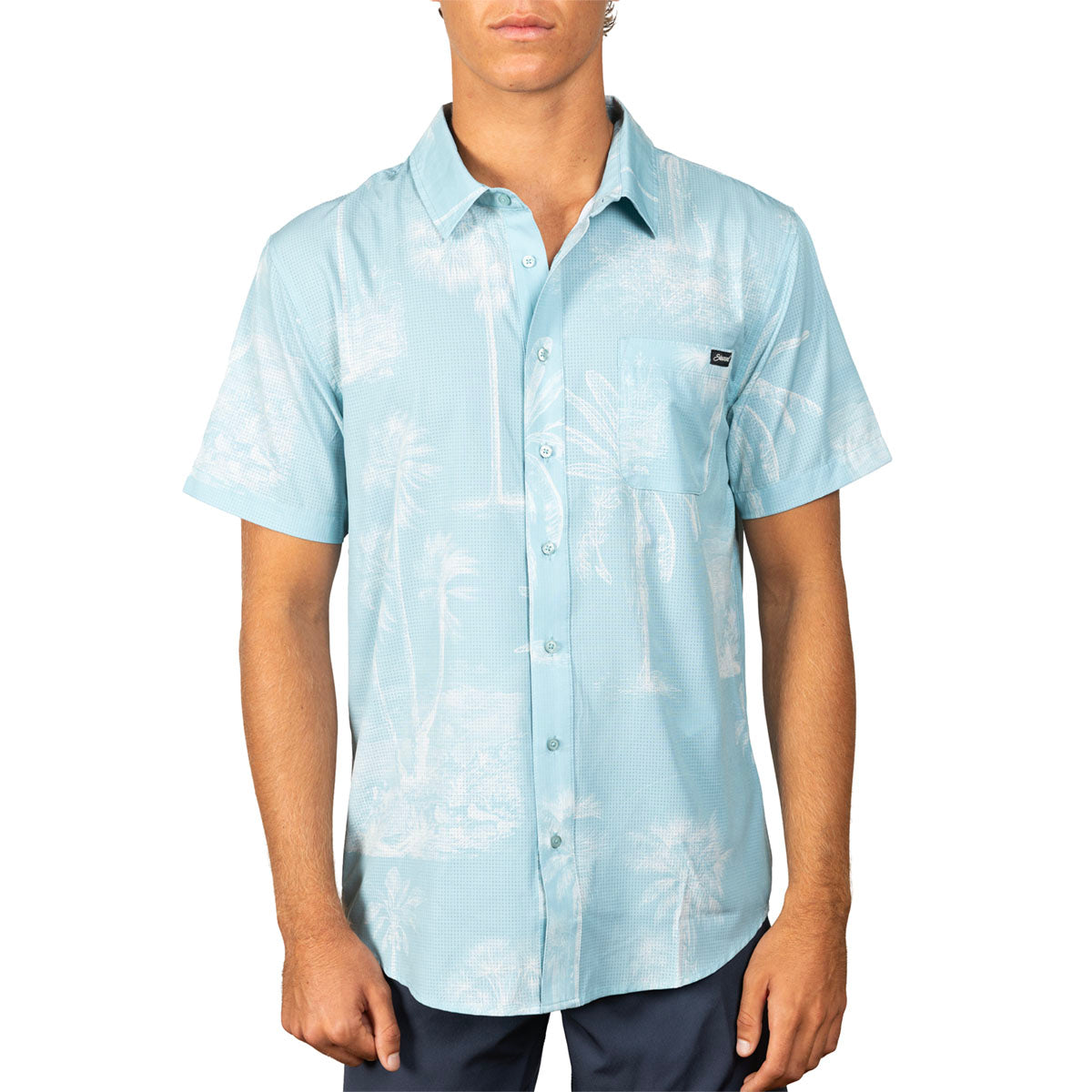A man wearing a light blue short-sleeve button-up shirt with a white palm tree pattern, paired with dark pants, against a plain white background.