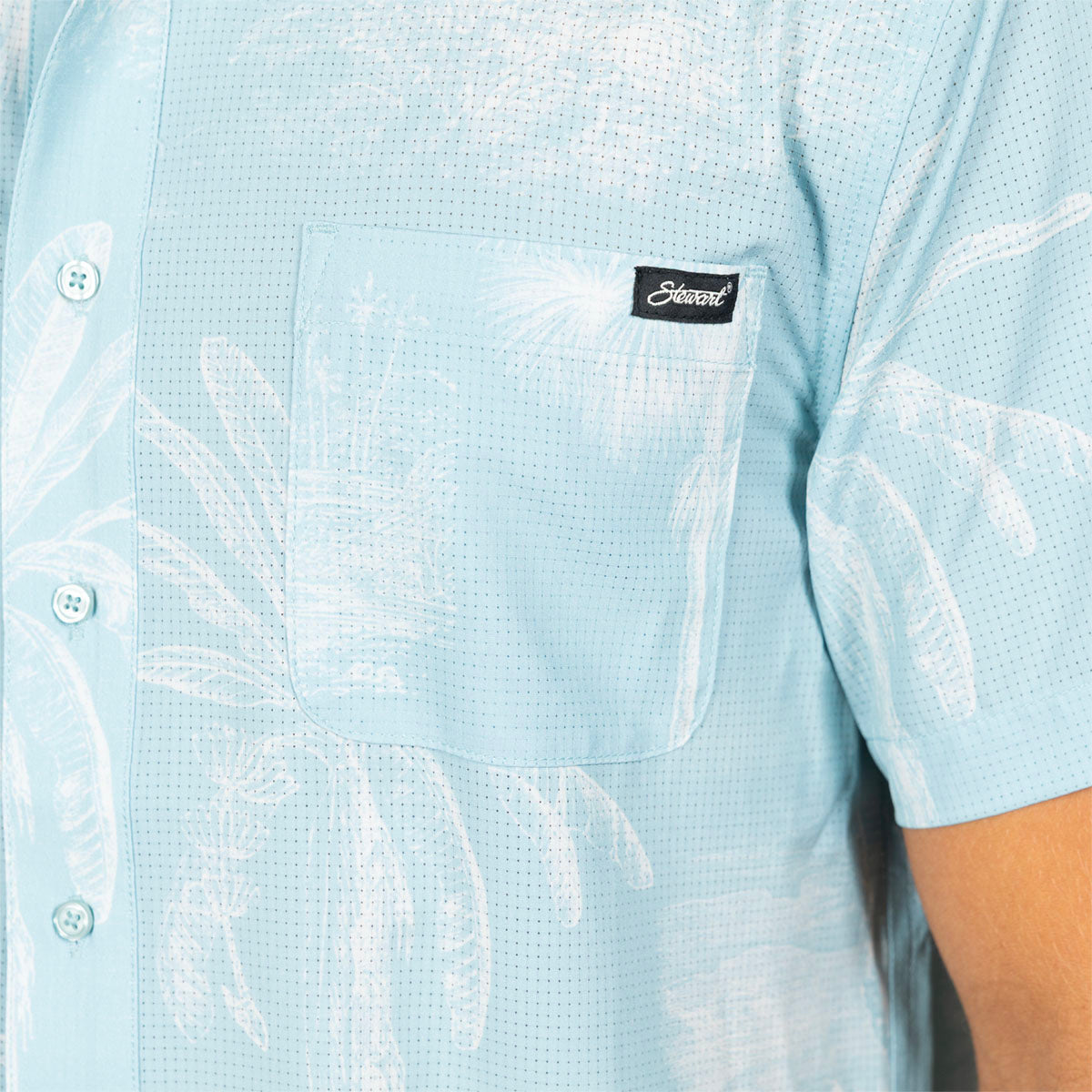 Close-up of a light blue short-sleeve button-up shirt with a white tropical leaf pattern and a black brand patch on the chest pocket.