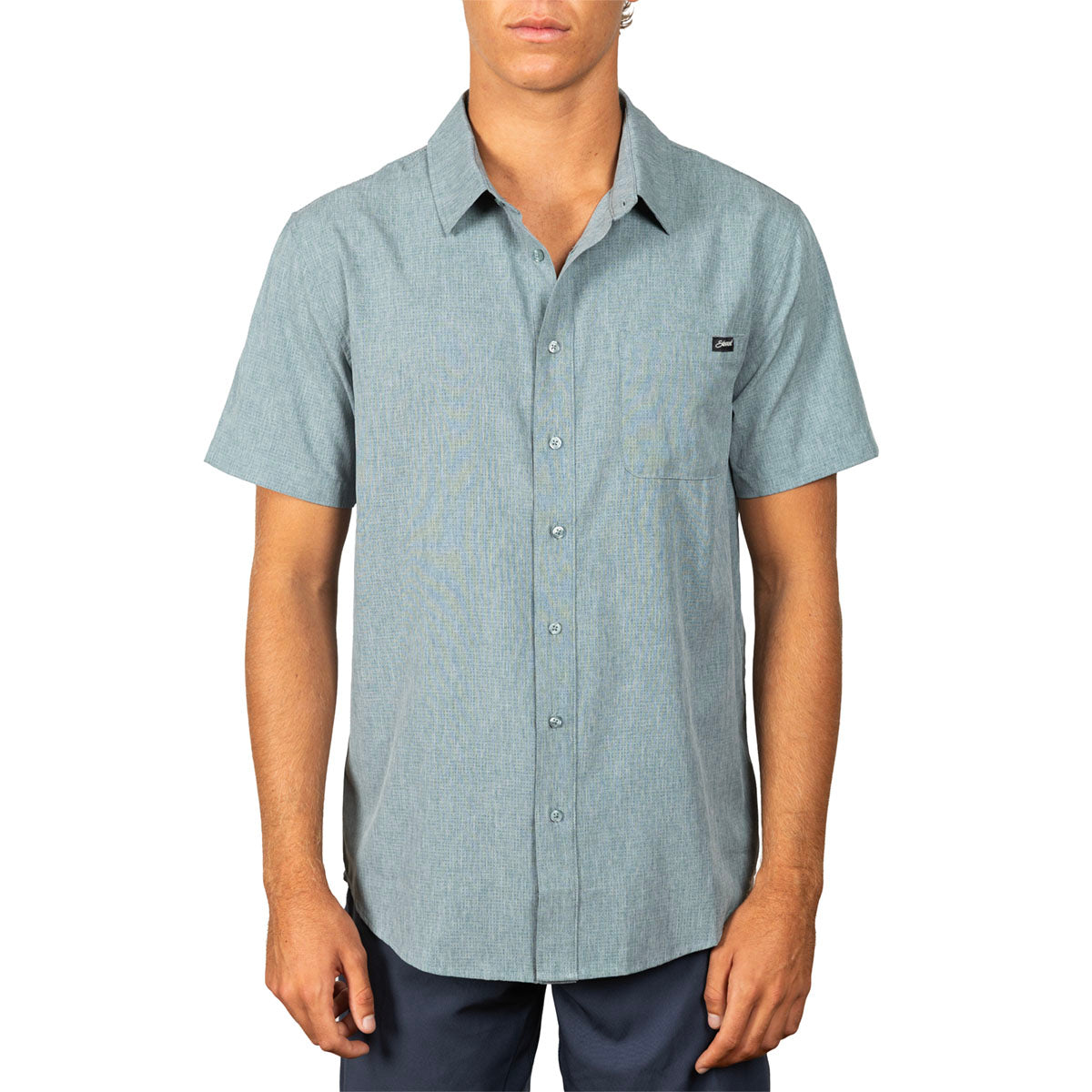 A man wearing a light blue short-sleeve button-up shirt with a small black logo patch on the left chest, paired with dark pants, against a plain white background.