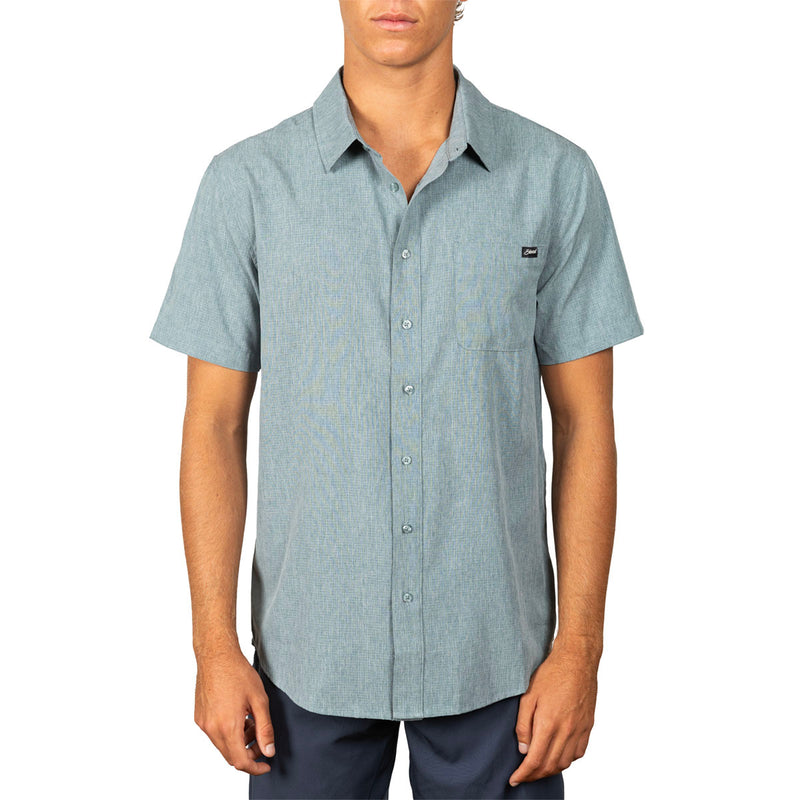 A man wearing a light blue short-sleeve button-up shirt with a small black logo patch on the left chest, paired with dark pants, against a plain white background.