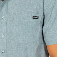 Close-up of a person wearing a light blue short-sleeve button-up shirt with a chest pocket and a black embroidered 'Stewart' logo patch.