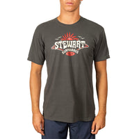 A man wearing a dark charcoal gray t-shirt with a red and white 'Stewart Surfboards' graphic on the chest, paired with navy blue shorts, against a plain white background.