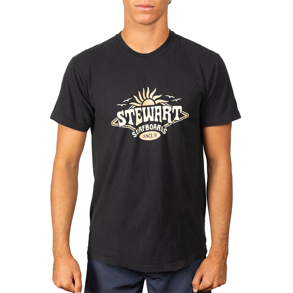 A man wearing a black short-sleeve t-shirt with a vintage-style beige graphic on the chest that reads 'STEWART SURFBOARDS SINCE 1972'.