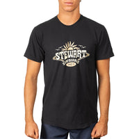 A man wearing a black short-sleeve t-shirt with a vintage-style beige graphic on the chest that reads 'STEWART SURFBOARDS SINCE 1972'.