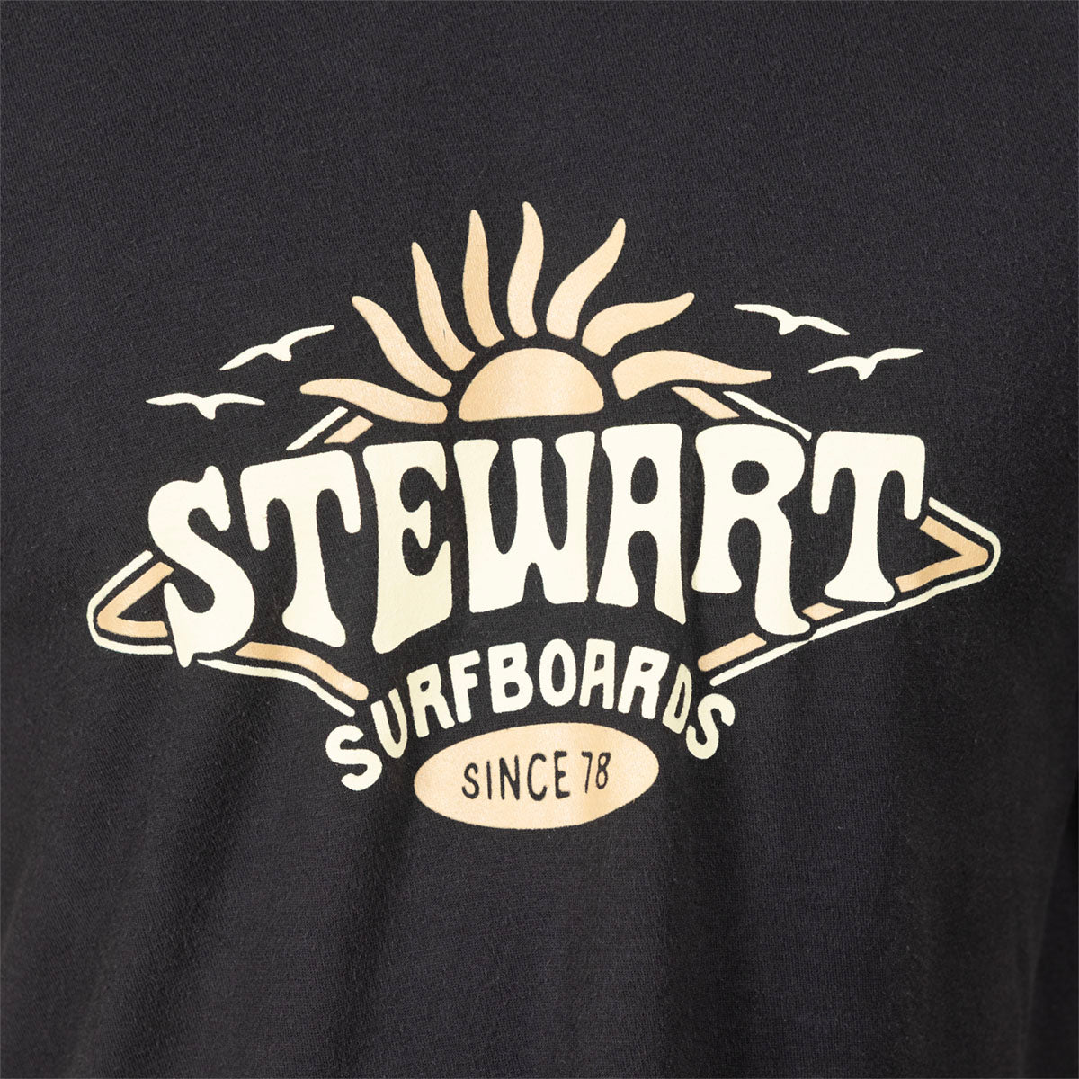 Close-up of a black T-shirt featuring a large vintage-style graphic with the words 'STEWART SURFBOARDS' and 'SINCE 78' in cream and beige tones, with a sun, waves, and birds above.