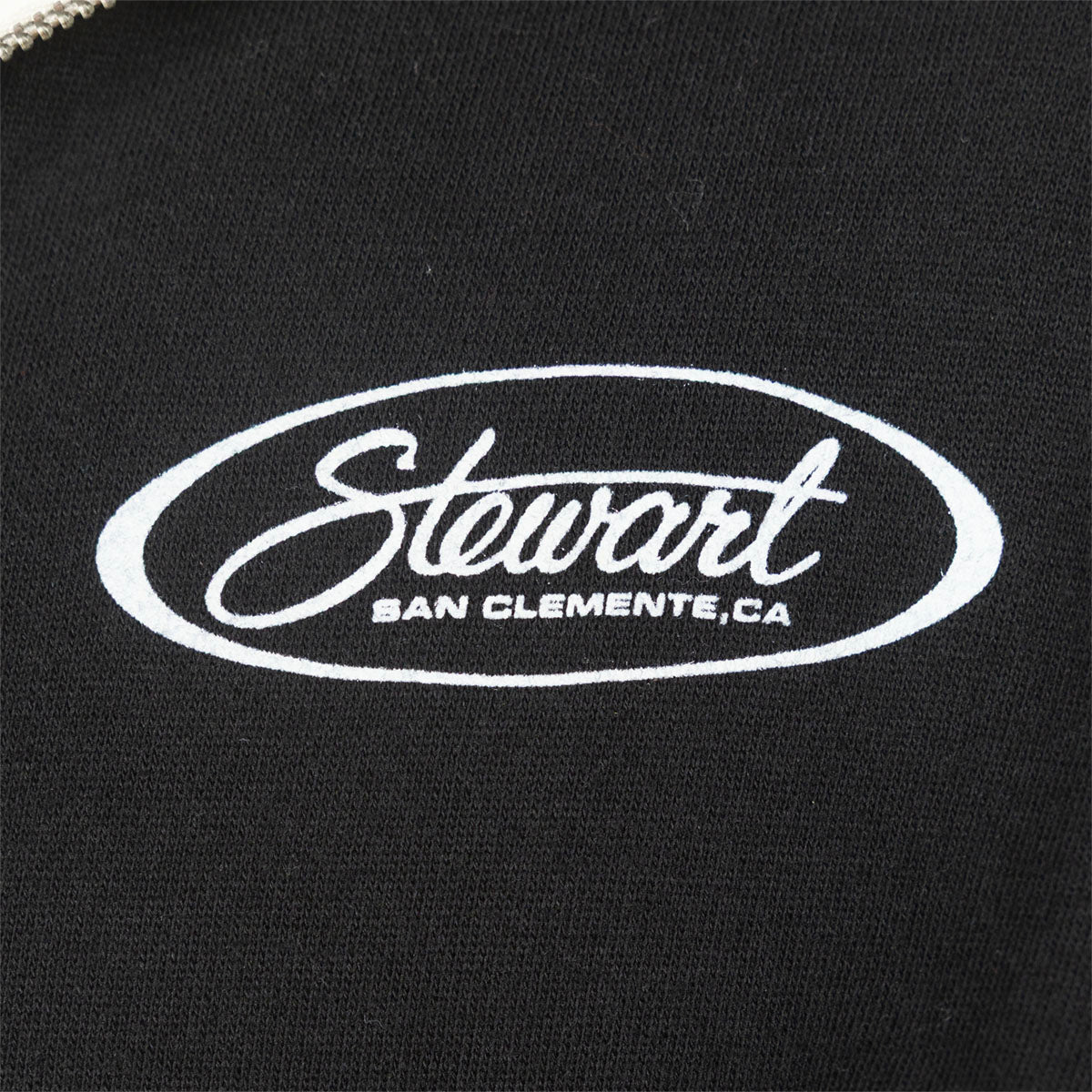 Close-up of a black fabric surface with a white embroidered oval logo reading 'Stewart' and smaller text 'SAN CLEMENTE,CA'.
