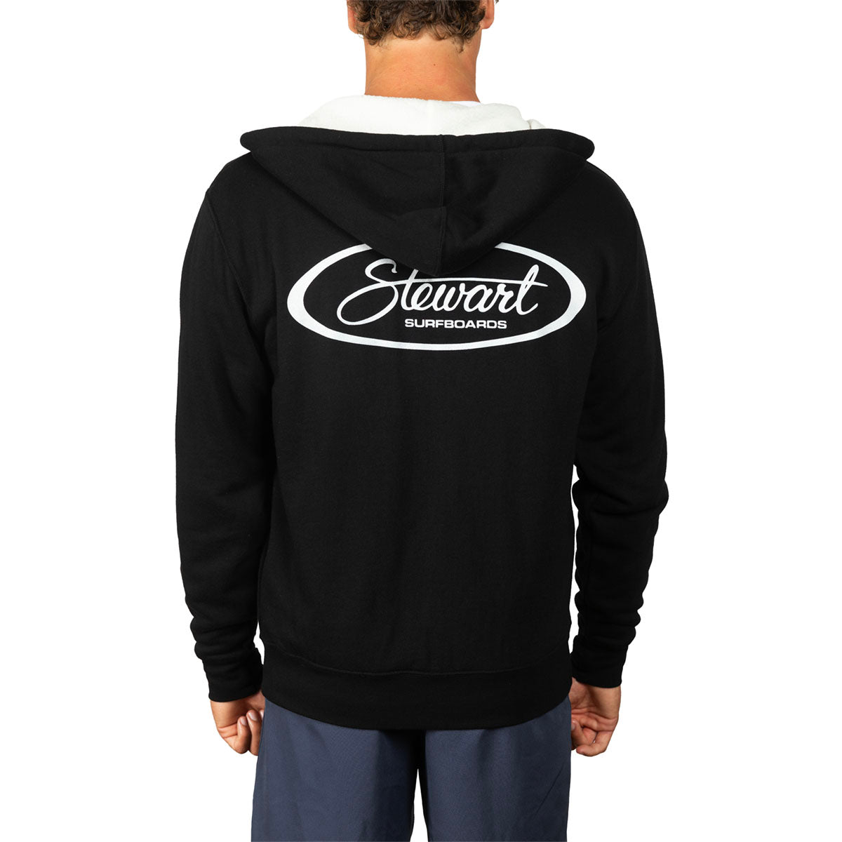 A person shown from the back wearing a black hooded sweatshirt with a large white Stewart Surfboards logo printed across the back, paired with blue pants, against a plain white studio background.