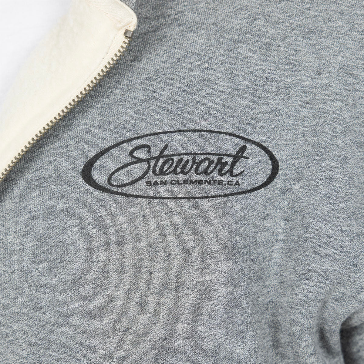 Close-up of a heather gray zip-up sweatshirt with a cream-colored fleece collar and a black embroidered oval logo reading 'Stewart San Clemente, CA' on the chest.