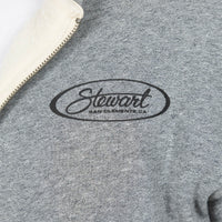 Close-up of a heather gray zip-up sweatshirt with a cream-colored fleece collar and a black embroidered oval logo reading 'Stewart San Clemente, CA' on the chest.