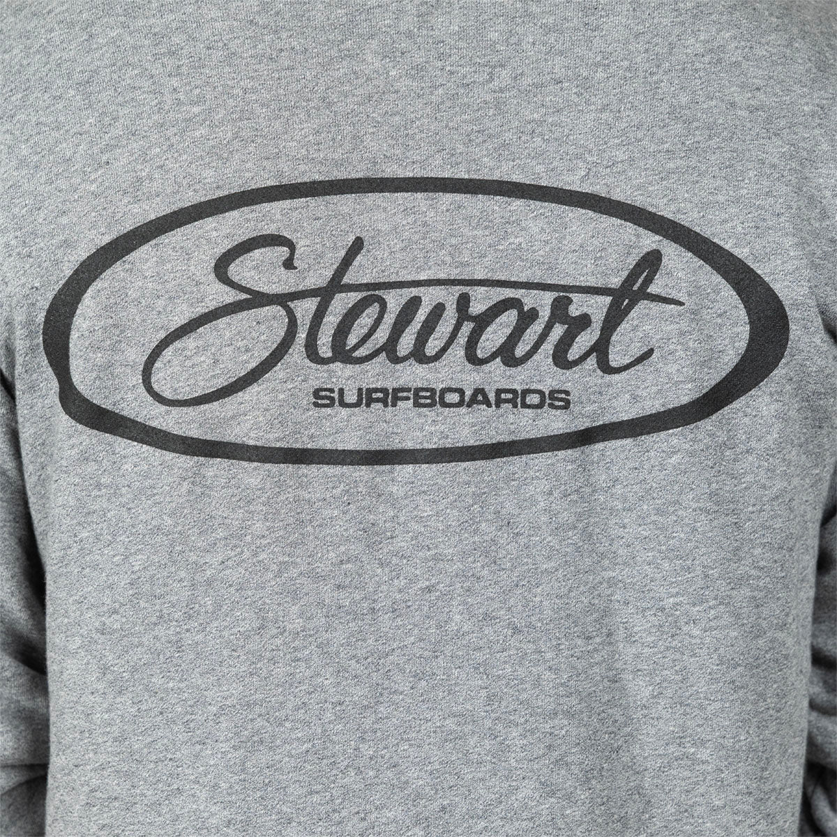 Close-up of a heather gray sweatshirt featuring a black oval graphic with the script word 'Stewart' and the text 'SURFBOARDS' underneath.