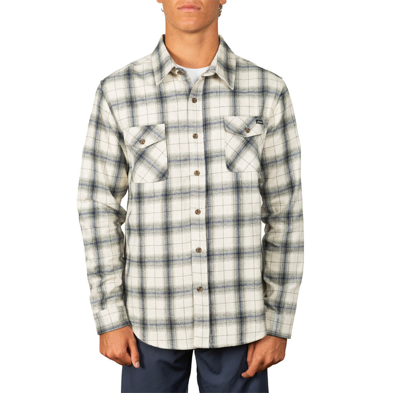 A man wearing a long-sleeve plaid button-up shirt in off-white with gray and navy lines, paired with dark blue pants, against a plain white background.