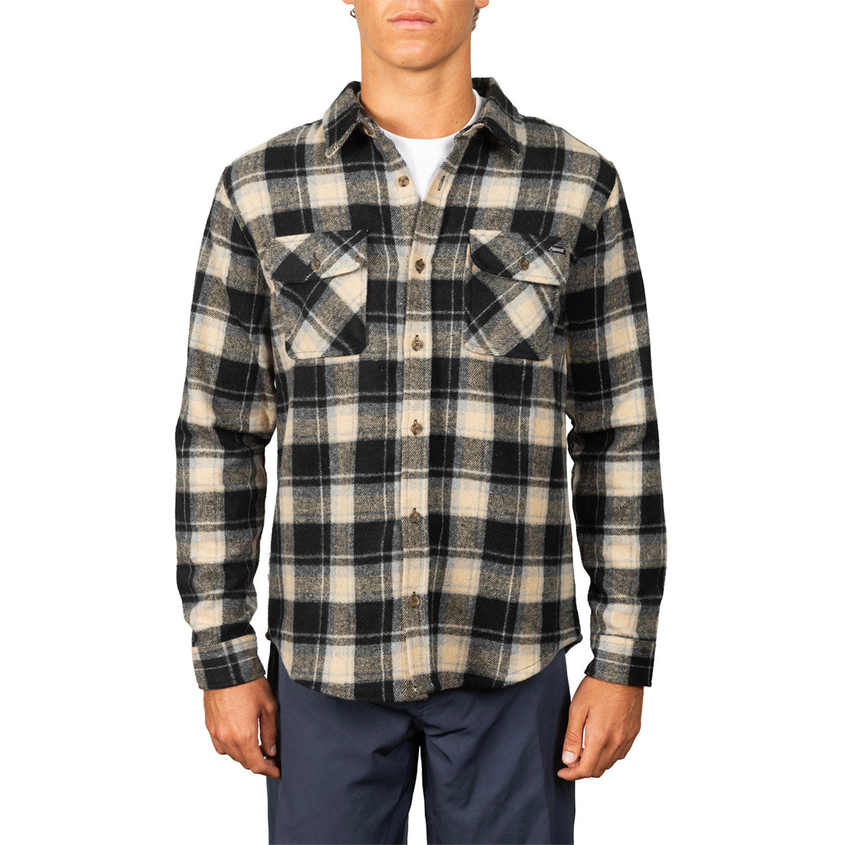 A man wearing a long-sleeve button-up plaid flannel shirt in black and beige, paired with navy pants, against a plain white background.