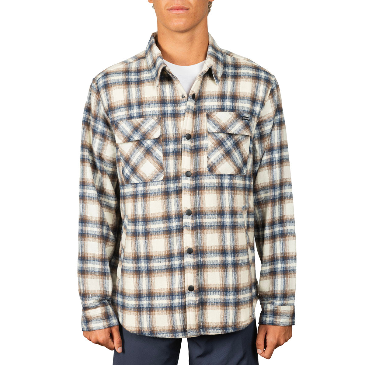 A man wearing a long-sleeve plaid button-up shirt in beige, brown, and blue, paired with a white t-shirt and navy pants, against a plain white background.
