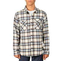 A man wearing a long-sleeve plaid button-up shirt in beige, brown, and blue, paired with a white t-shirt and navy pants, against a plain white background.