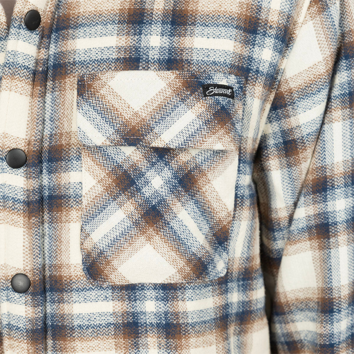 Close-up of a plaid button-front shirt jacket with a chest pocket. The fabric has a textured herringbone weave in cream, blue, and brown, with a small black brand tab reading 'Stewart' on the pocket.