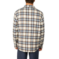 A man viewed from the back wearing a long-sleeve plaid flannel shirt in cream, brown, and blue, paired with navy pants, against a plain white background.
