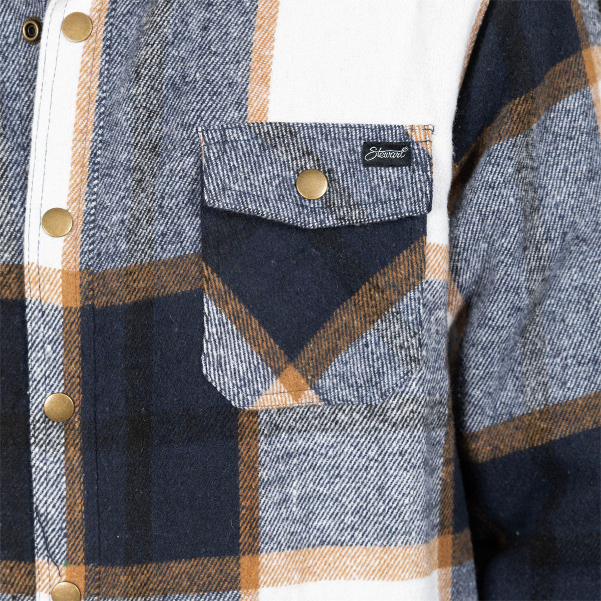 Close-up of a plaid flannel shirt pocket with a snap button flap and a visible Stüssy label, showing navy, gray, tan, and white checks.