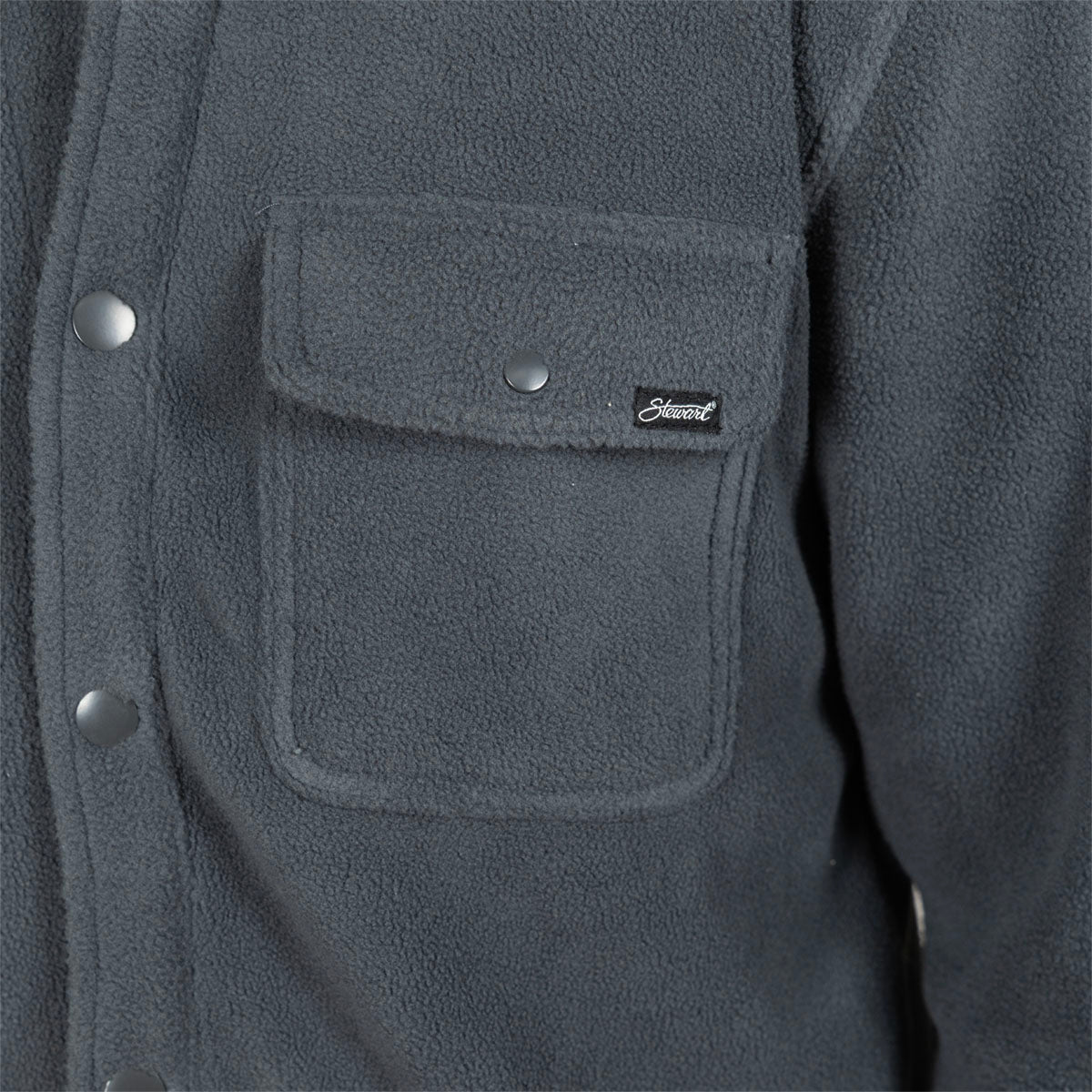Close-up of a dark gray fleece jacket featuring a chest pocket with a flap, silver snap buttons, and a small black brand patch.