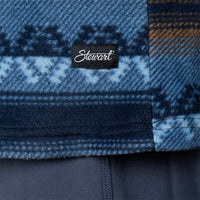 Close-up of a blue patterned textile with a black fabric label reading Stewart in white script, shown against a dark navy fabric background.