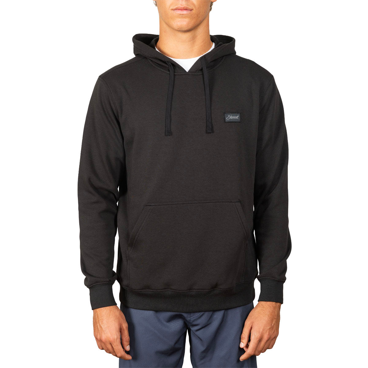 A man wearing a black pullover hoodie with a small rectangular chest logo patch, paired with blue pants, shown against a plain white background.