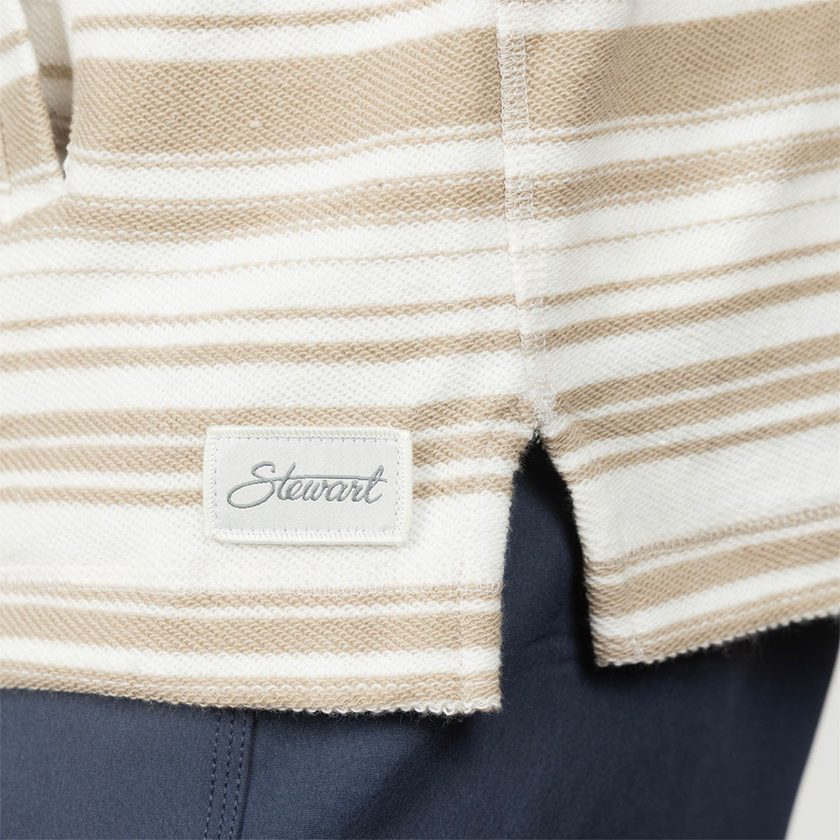 Close-up of a striped woven garment with a small sewn label reading 'Stewart' in script, showing beige and off-white horizontal stripes and a dark navy fabric below.