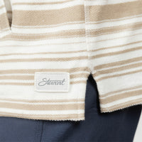 Close-up of a striped woven garment with a small sewn label reading 'Stewart' in script, showing beige and off-white horizontal stripes and a dark navy fabric below.