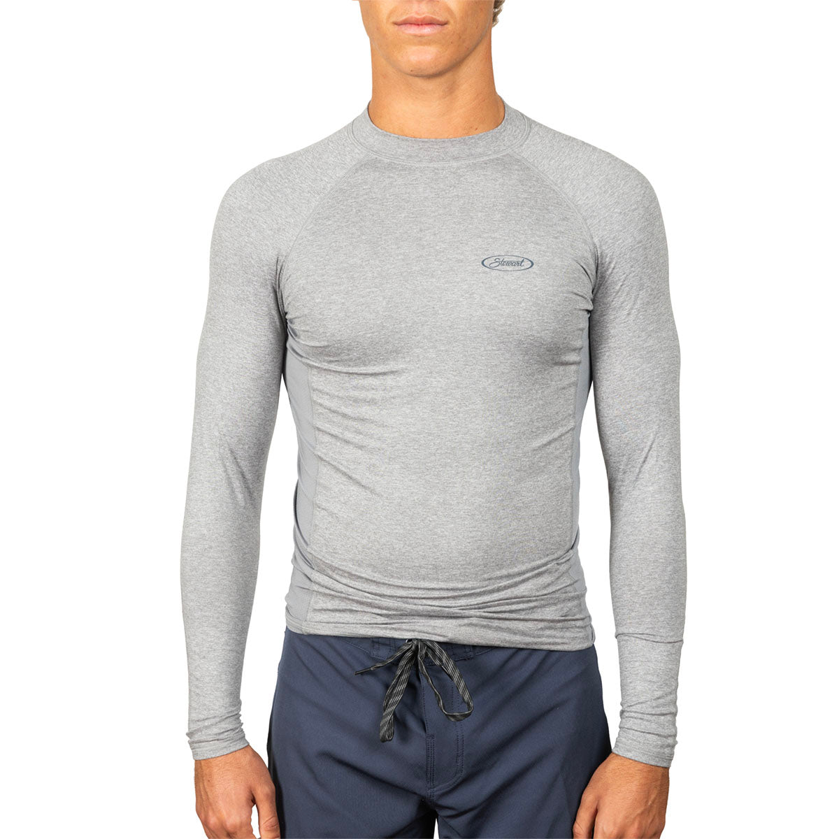 A man wearing a light gray long-sleeve compression shirt with a small logo on the left chest, paired with navy blue drawstring shorts, against a plain white background.