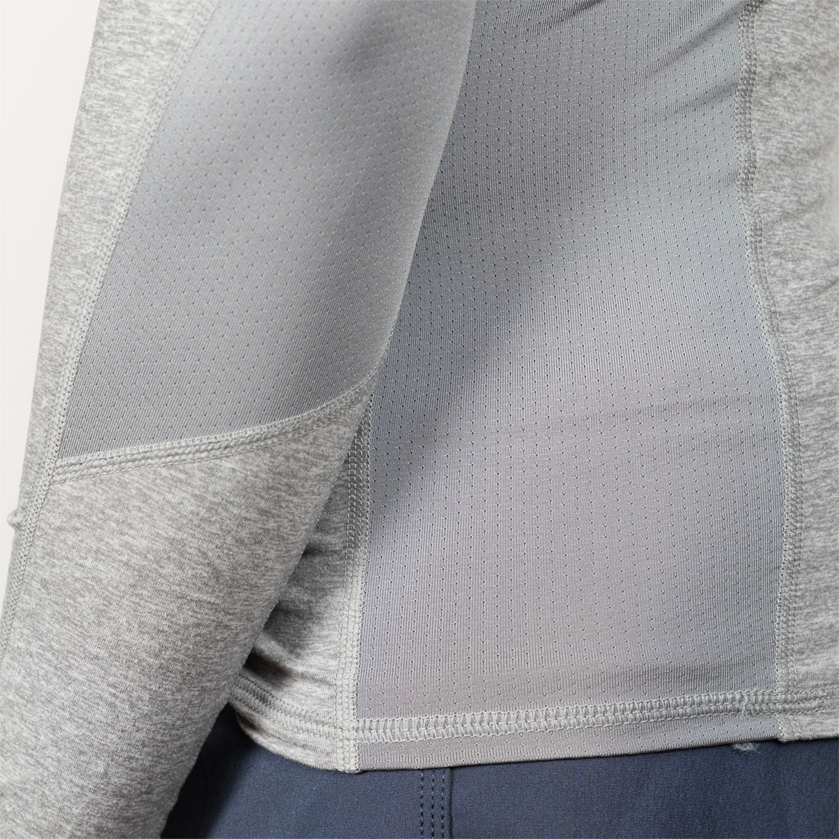 Close-up of a light gray athletic long-sleeve top with textured mesh and heathered fabric panels, shown on a model wearing dark blue bottoms.