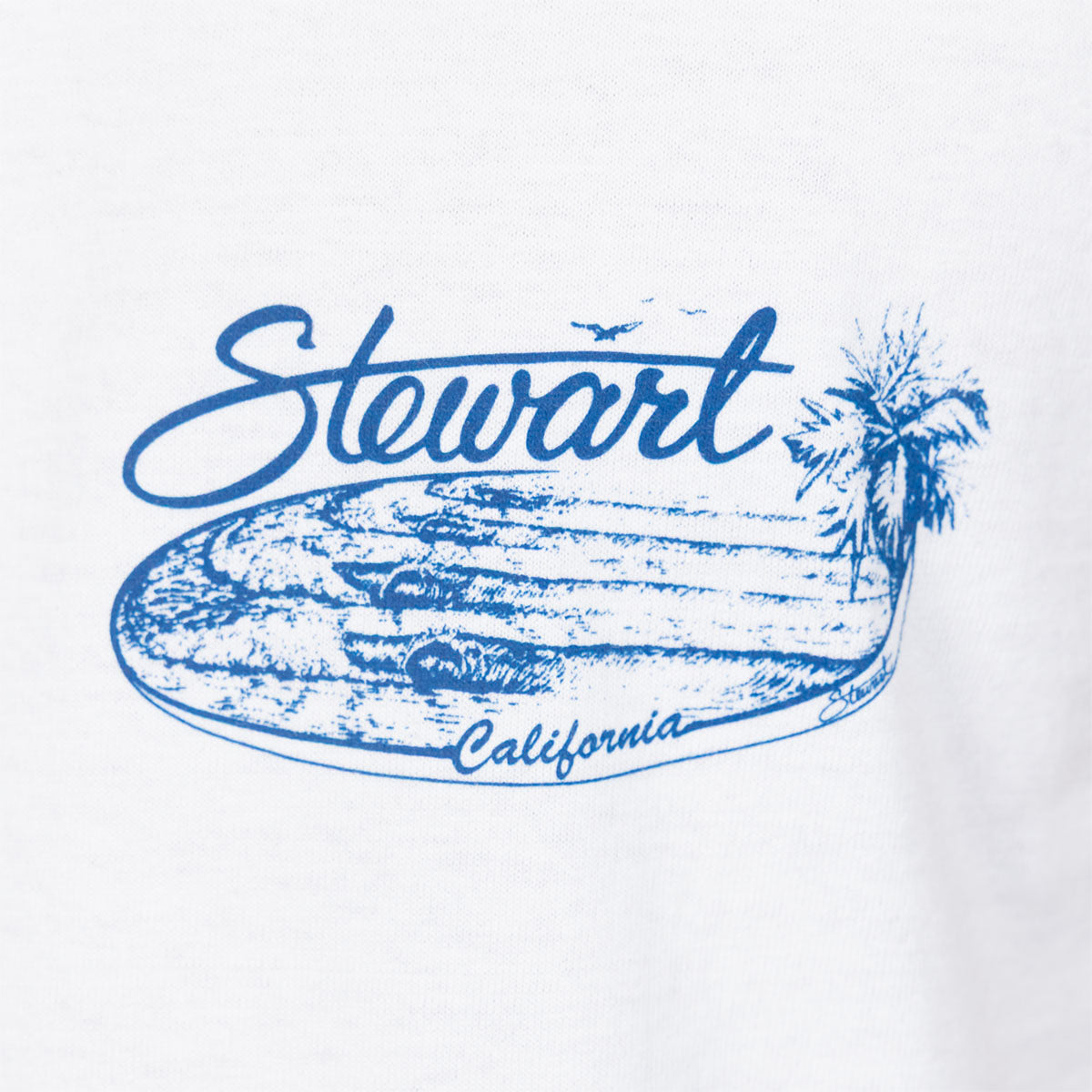 A close-up of a white fabric surface featuring a blue graphic print with the word 'Stewart' in script, a palm tree, a surfboard illustration, and the word 'California' along the bottom.